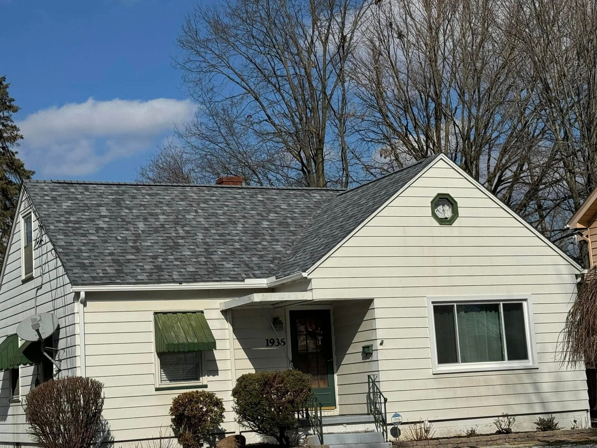 Small white house with gray roof, green awning, front steps, and bare trees under a blue sky.