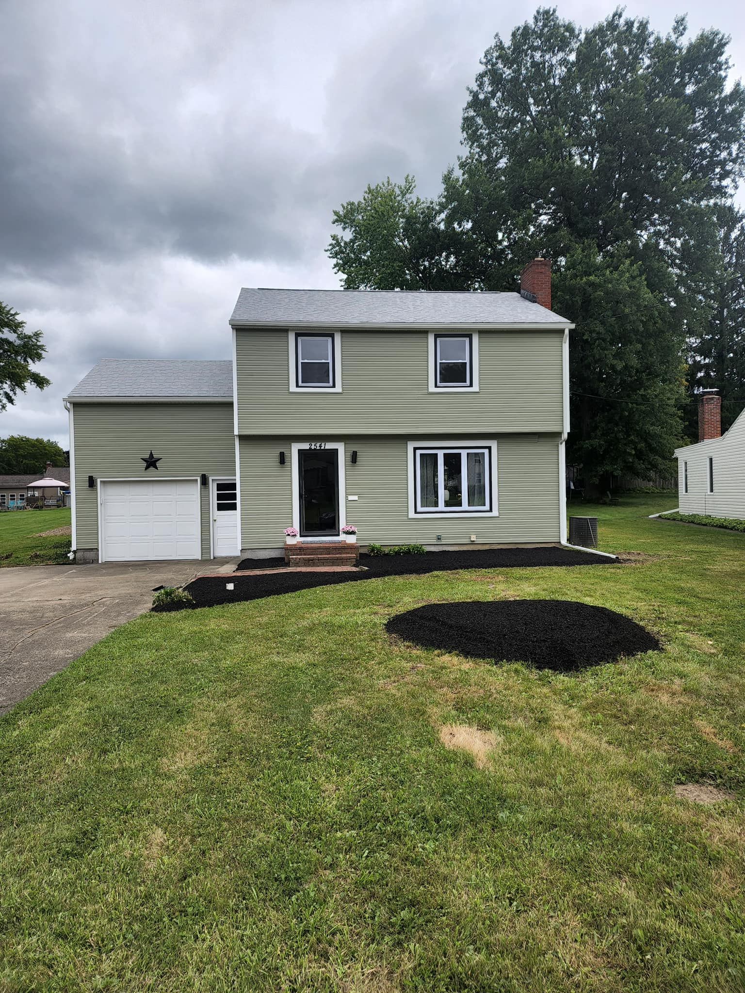 Two-story green house with attached garage, front porch, and gravel driveway under cloudy skies