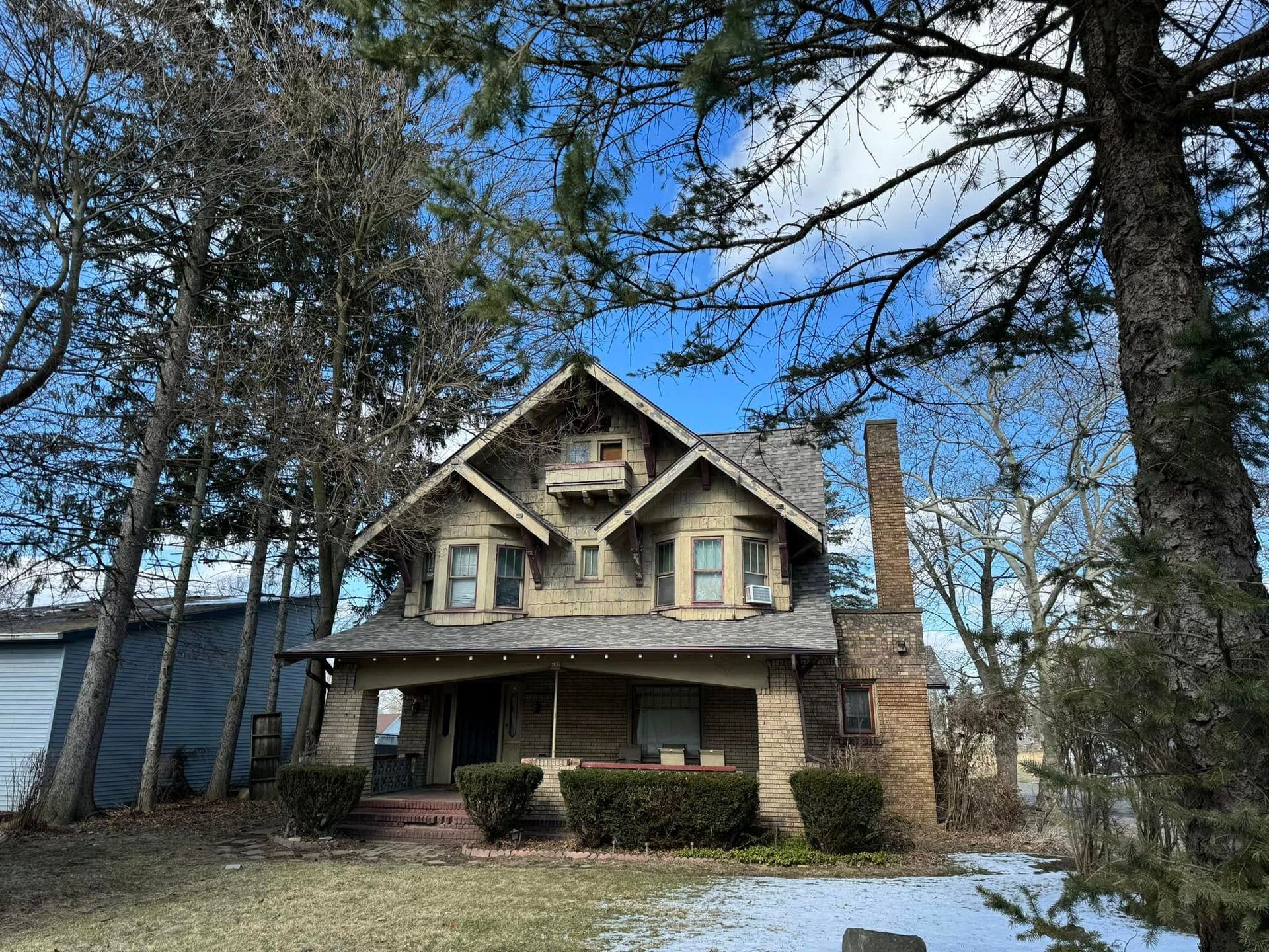 Two-story house with a front porch, stone chimney, and bare trees under a blue sky