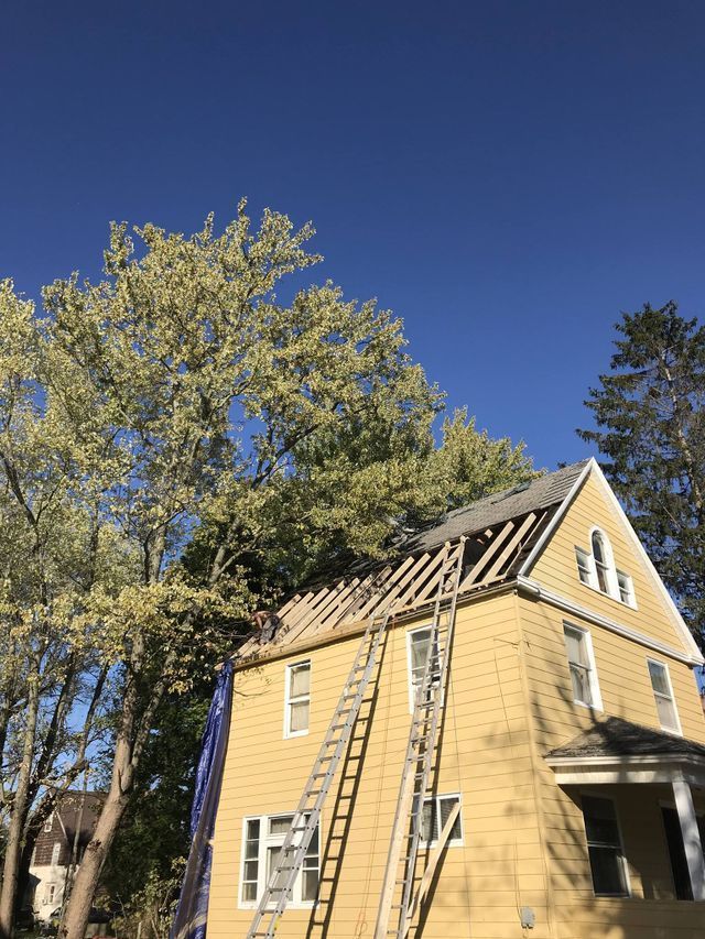 Yellow house under repair with exposed roof framing beside tall trees against a clear blue sky