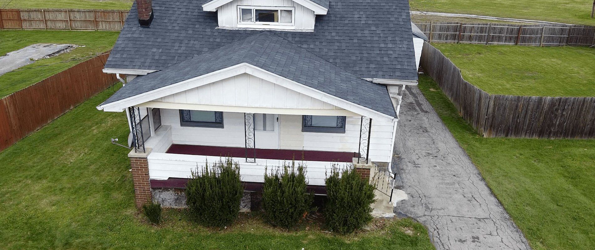 White house with gray roof, front porch, and a gravel driveway beside green grass and hedges