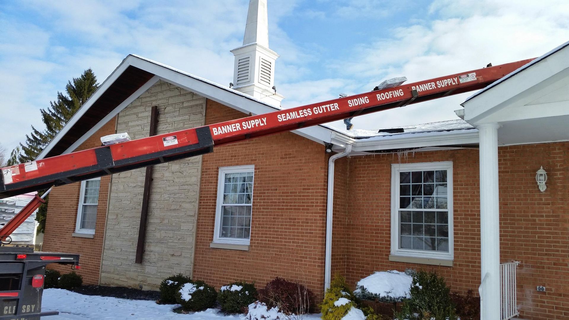 Snow-covered brick building with a red ladder propped across the roof in front of a church steeple