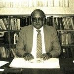 A man in a suit and tie is sitting at a desk in front of a bookshelf.