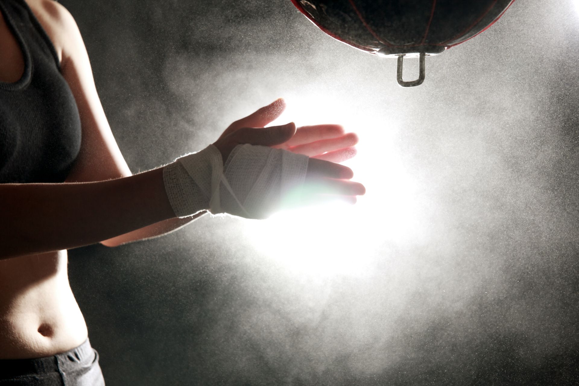 Donna che batte le mani con i polsi fasciati davanti a un sacco da boxe; polvere nell'aria.