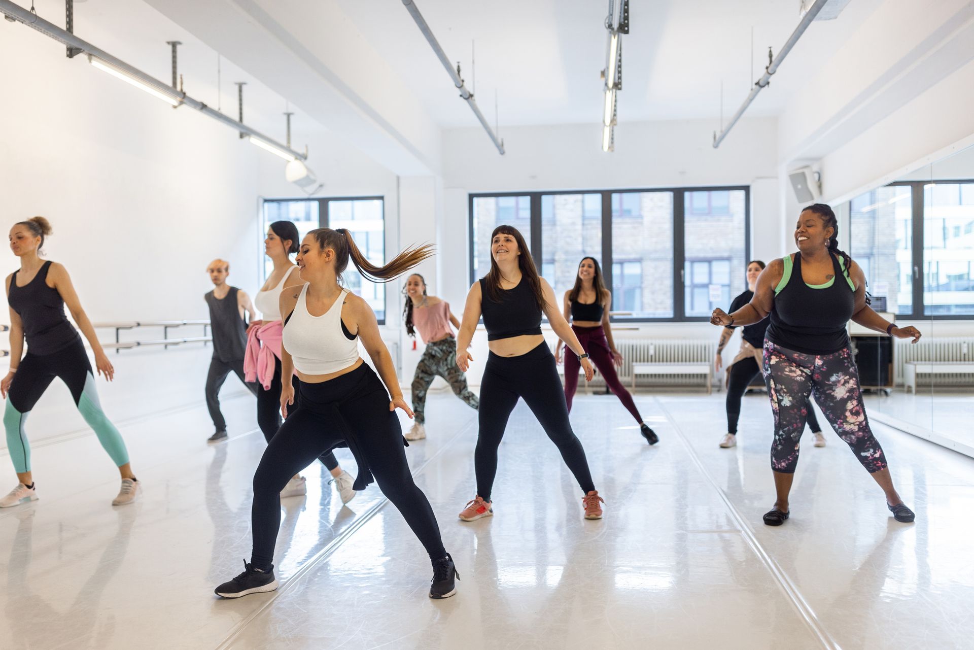 Persone a lezione di danza, studio con parete a specchio. Si appoggiano, si allungano e ballano. Spazio luminoso con luce.