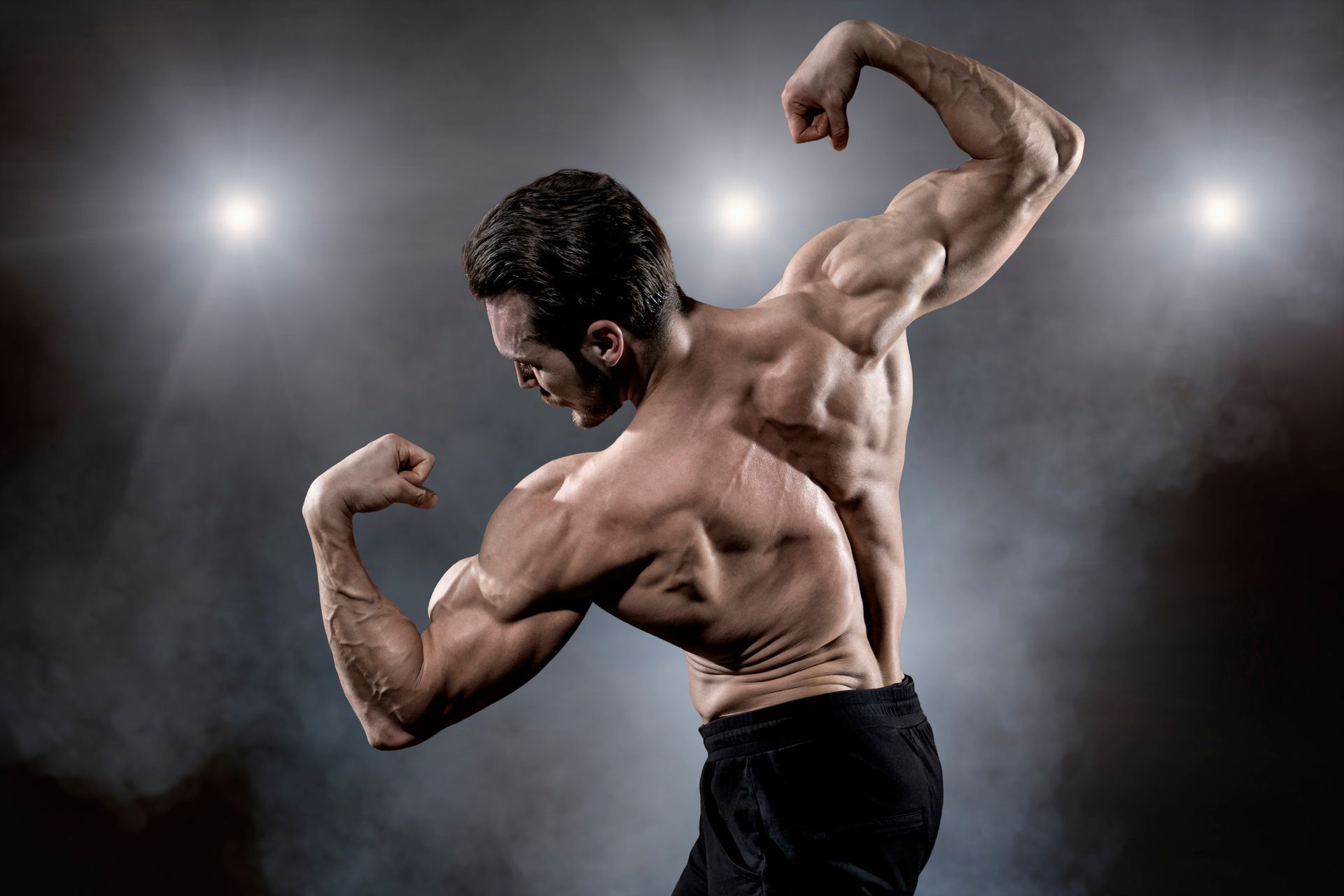 Uomo muscoloso che flette i bicipiti in un ambiente buio, sotto luci intense.