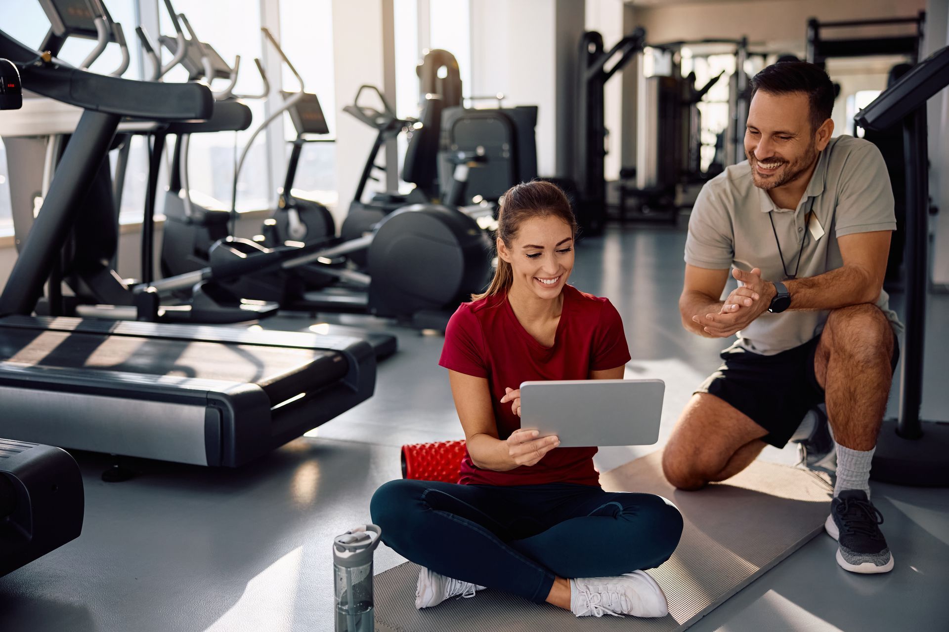 Donna e uomo in palestra, guardano insieme un tablet, sorridenti. Sullo sfondo si vedono gli attrezzi da fitness.
