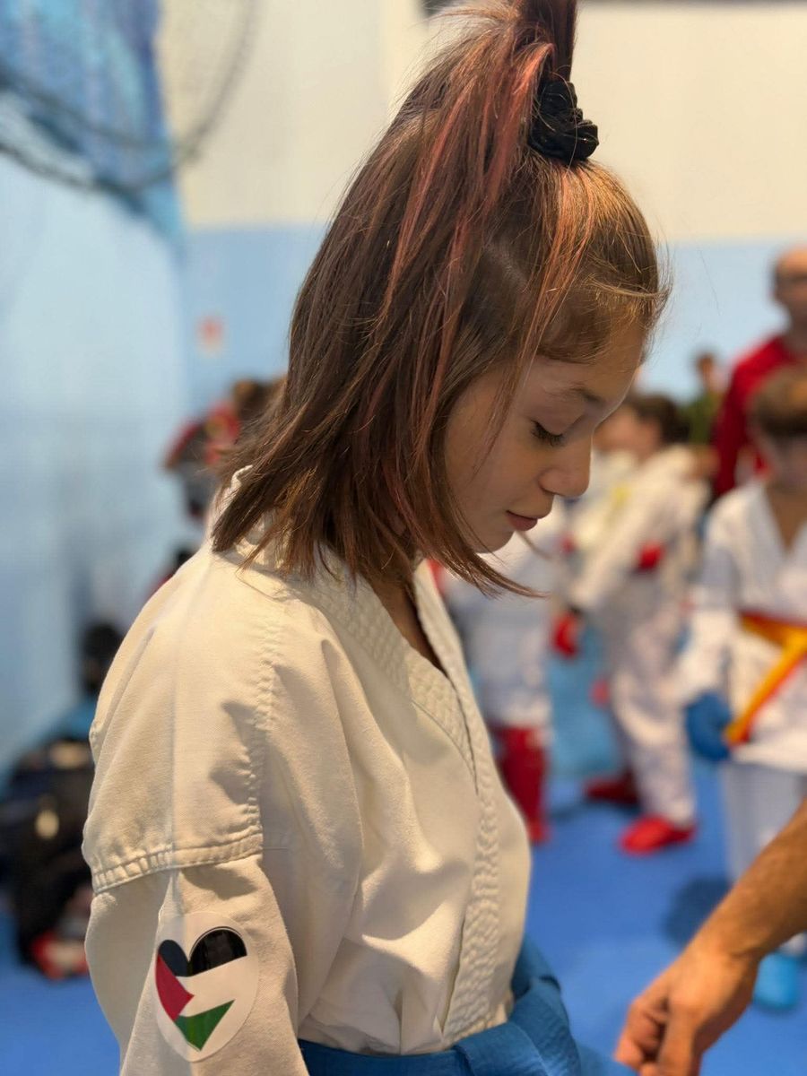 Giovane in uniforme bianca da arti marziali, con lo sguardo rivolto verso il basso. Stemma con bandiera palestinese sulla manica. Ambientazione interna.