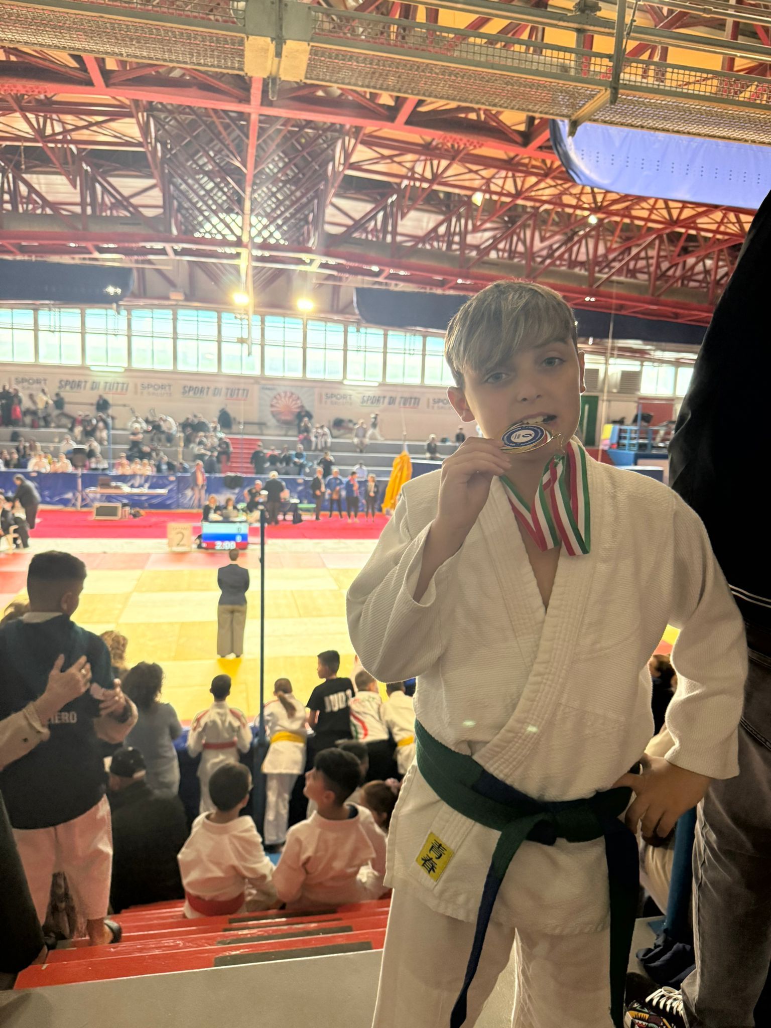 Ragazzo in uniforme da judo con medaglia, che la morde, al chiuso durante una gara.