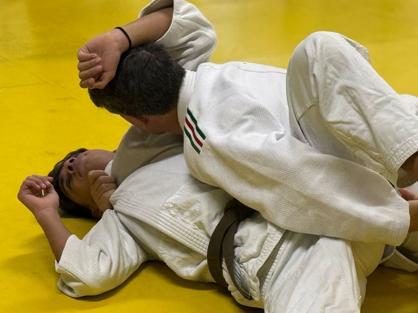 Due persone in uniforme da judo si esercitano su un materassino giallo. Una è sopra e controlla le braccia dell'altra.