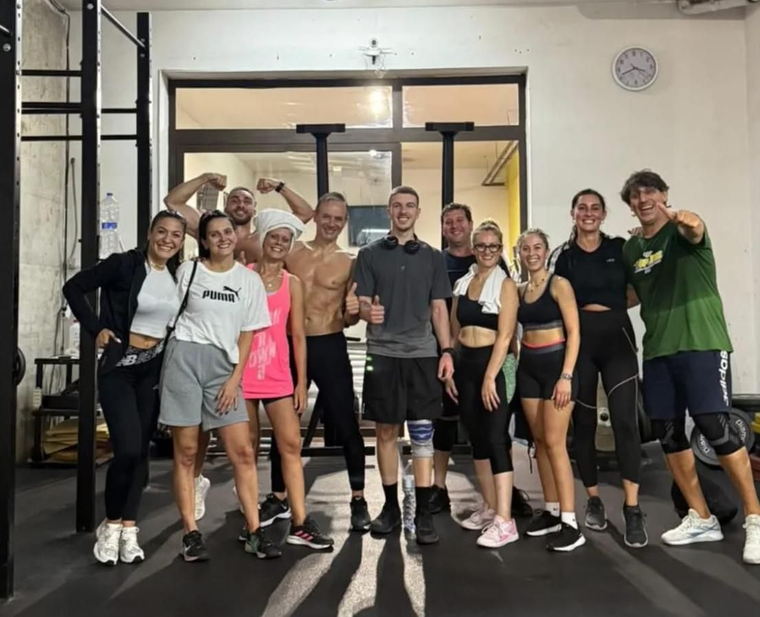 Un gruppo di persone posa insieme in una palestra. Alcuni si flettono, altri sorridono, sotto luci intense.