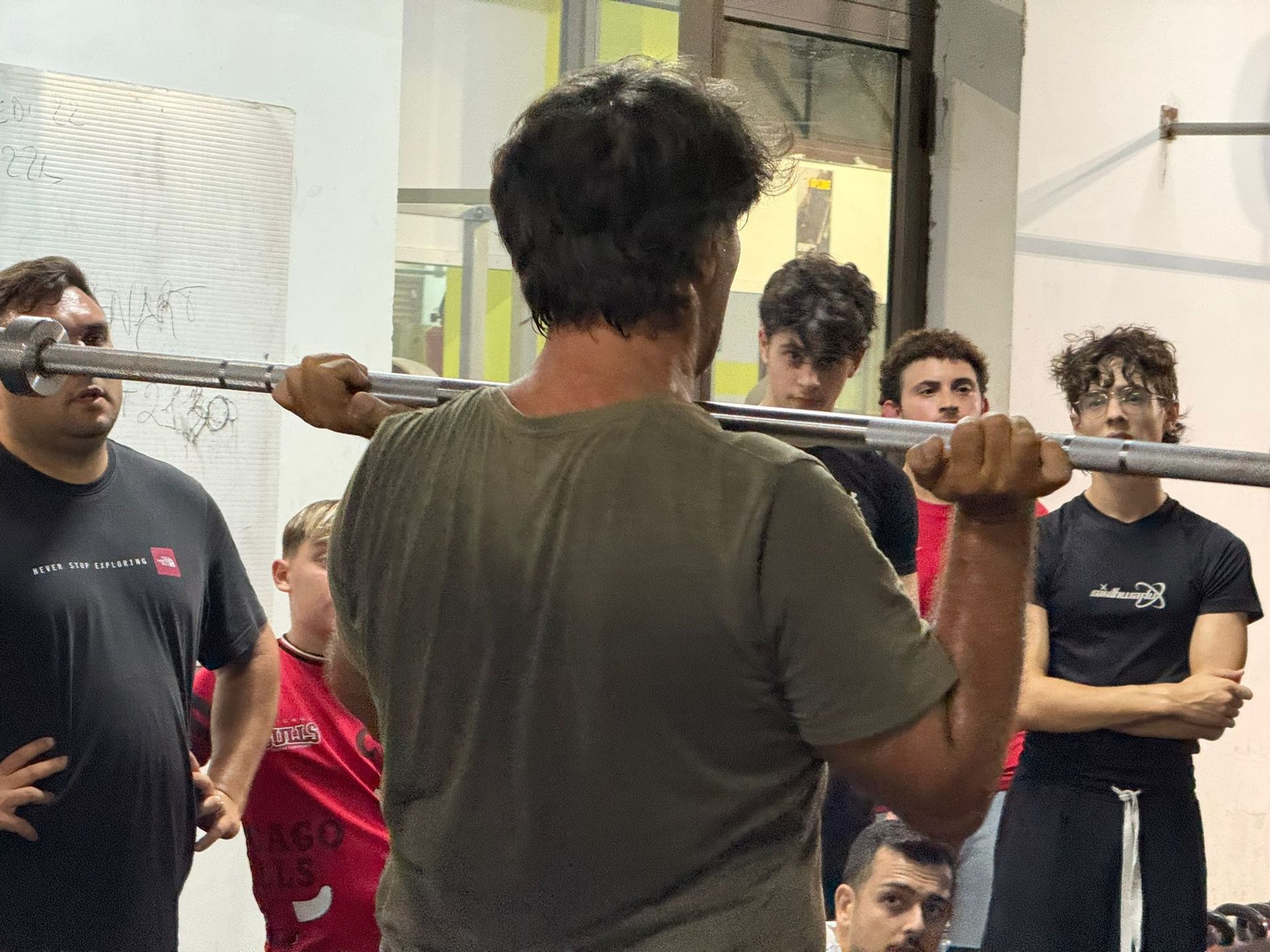 Un uomo tiene in mano un bilanciere e mostra come sollevarlo a un gruppo di persone in palestra.