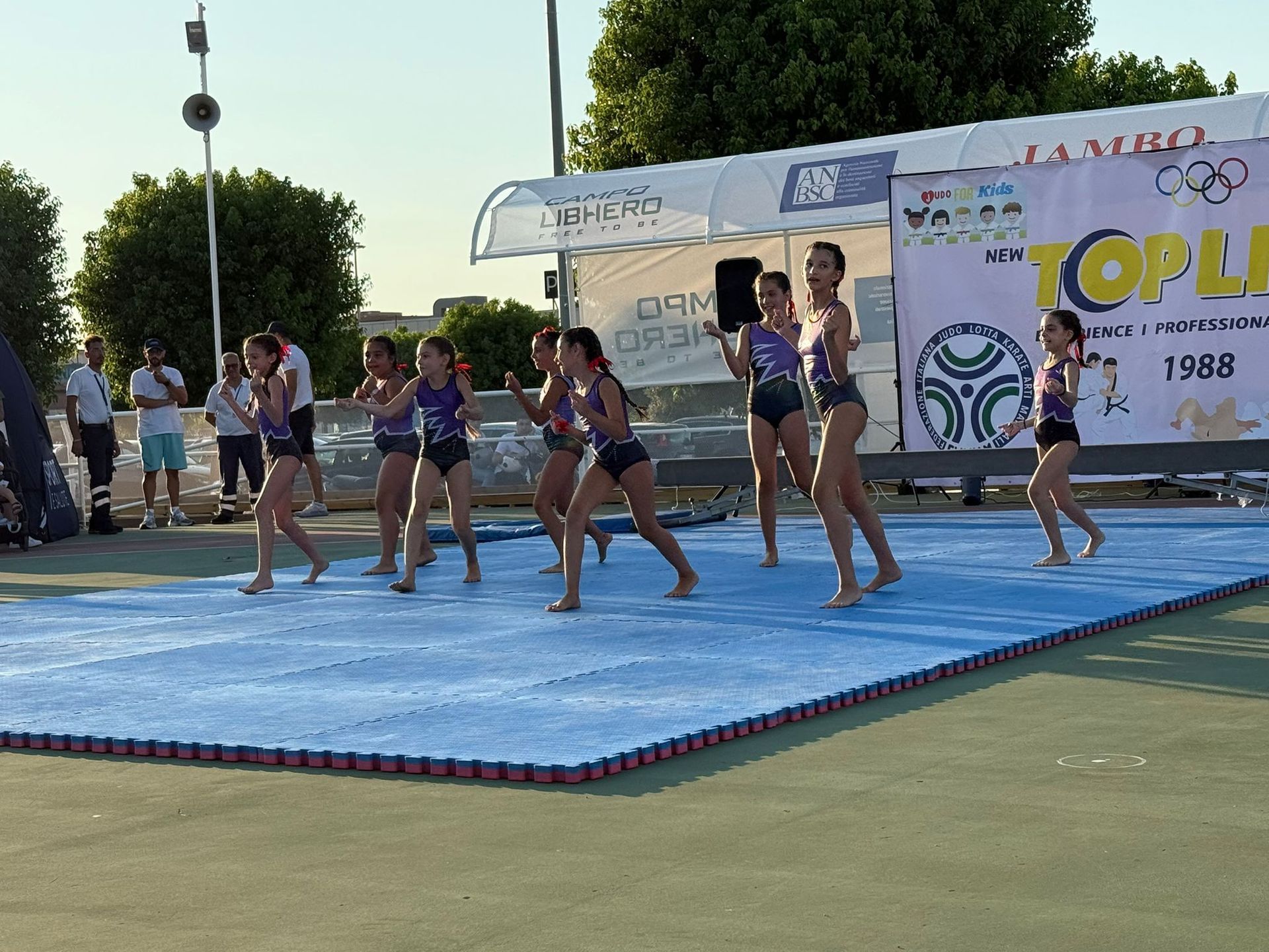 Un gruppo di giovani atleti in abbigliamento sportivo si esibisce su un tappeto blu all'aperto, davanti a un palco.