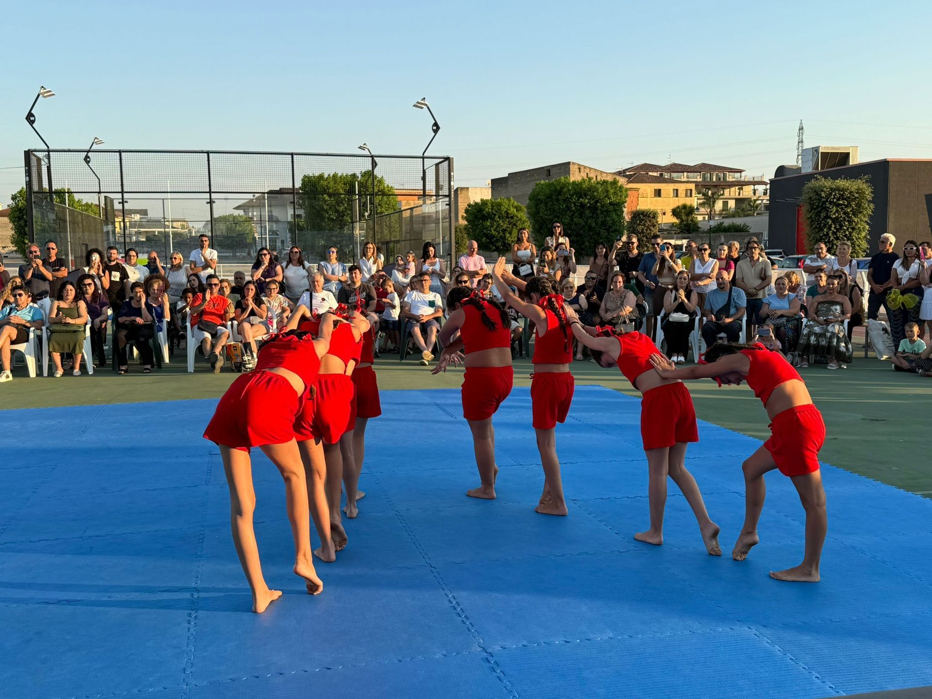 I ballerini in costume rosso si esibiscono all'aperto su un tappeto blu, piegandosi in avanti. Il pubblico osserva sullo sfondo.