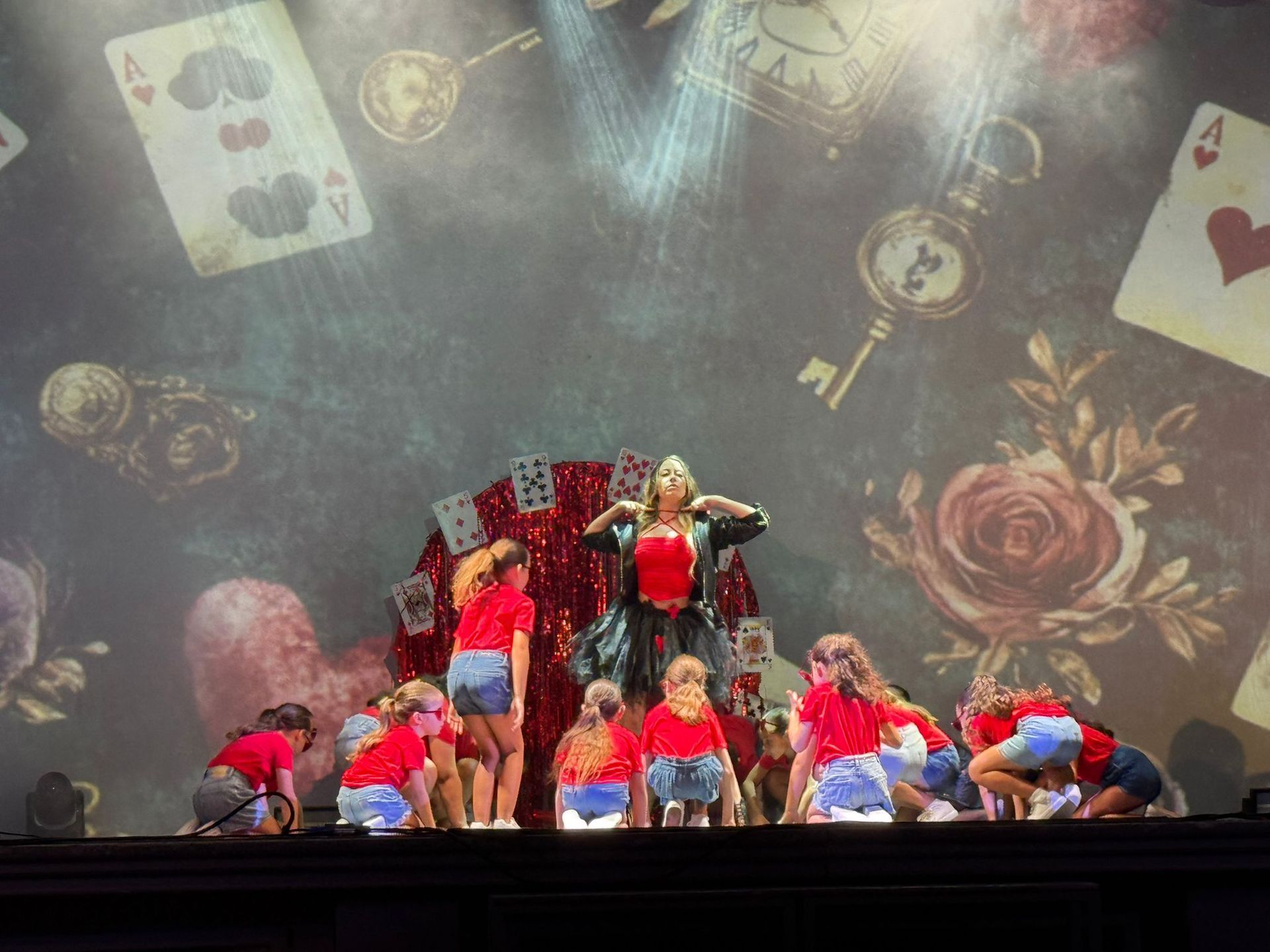 Uno spettacolo teatrale con ballerini in camicia rossa e pantaloncini di jeans, con una figura centrale in corsetto e giacca. Lo sfondo è a tema Alice nel Paese delle Meraviglie.