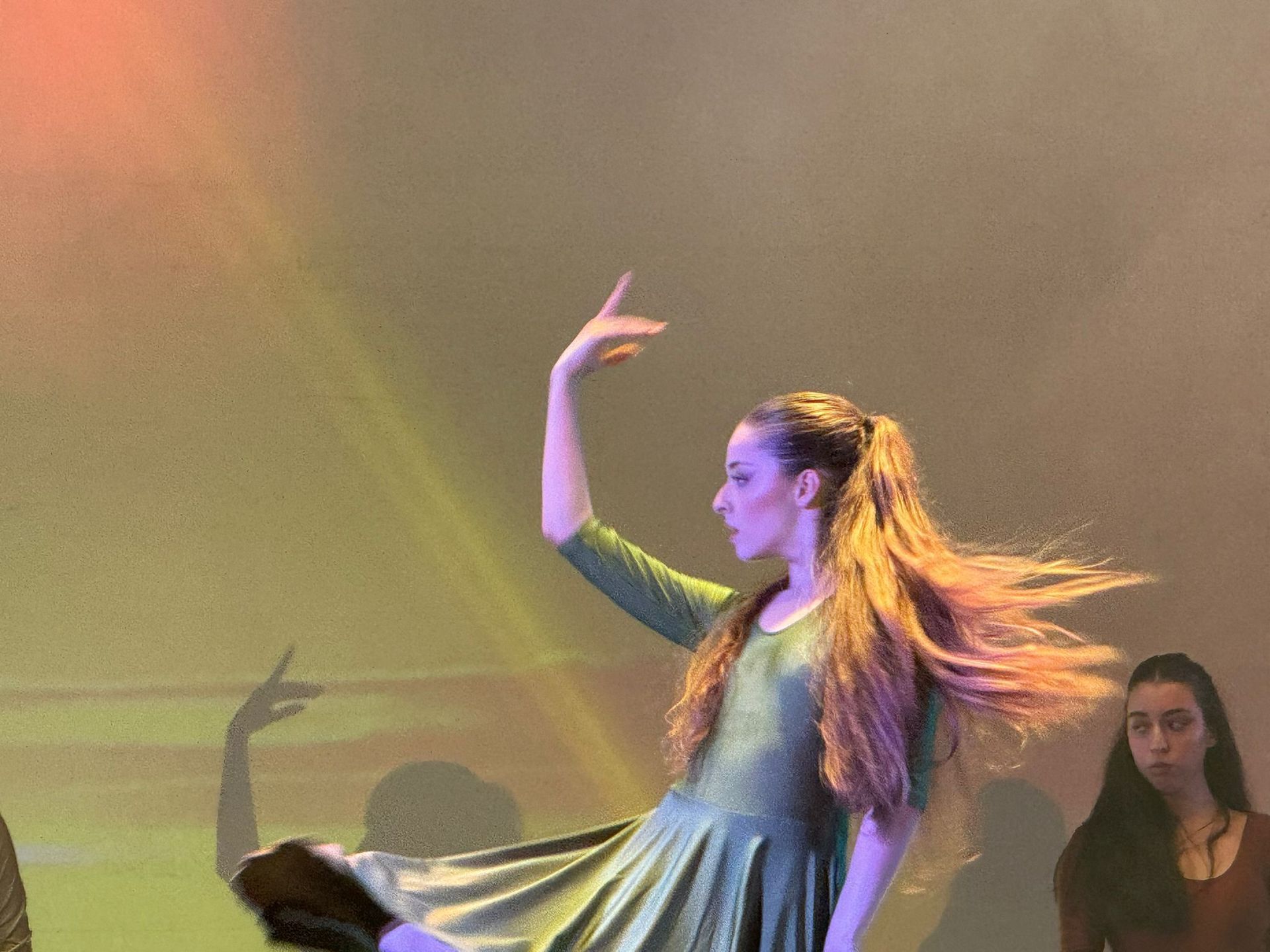 Donna in abito verde che balla sul palco, braccio alzato, capelli fluenti, ombra visibile.