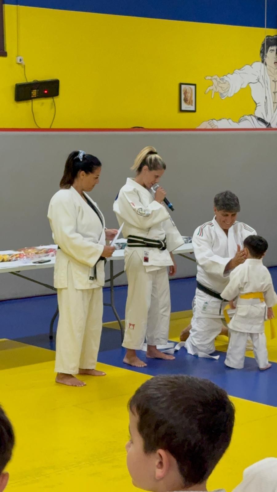 Cerimonia della cintura di judo: una persona in gi bianco riceve la cintura dagli istruttori. I bambini guardano, alcuni indossano gi gialli.