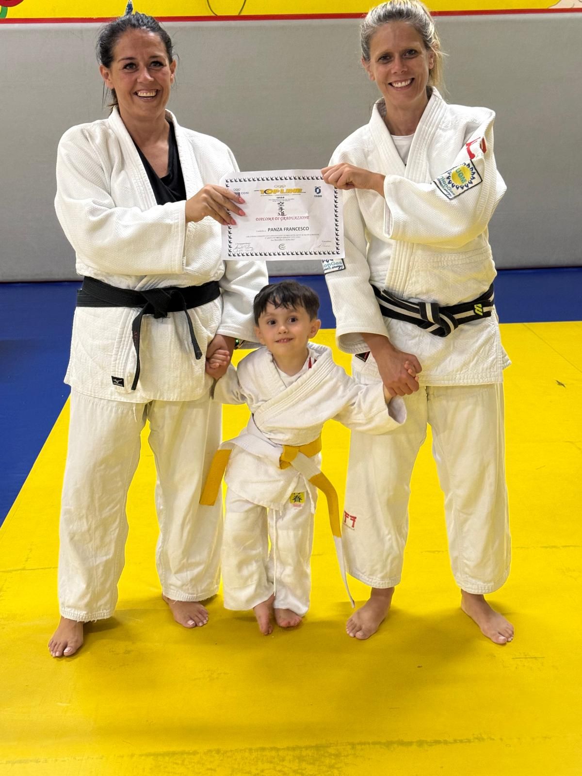 Due adulti in uniforme da judo affiancano un bambino in uniforme gialla da judo, con in mano un certificato. Sono su un tatami da judo.