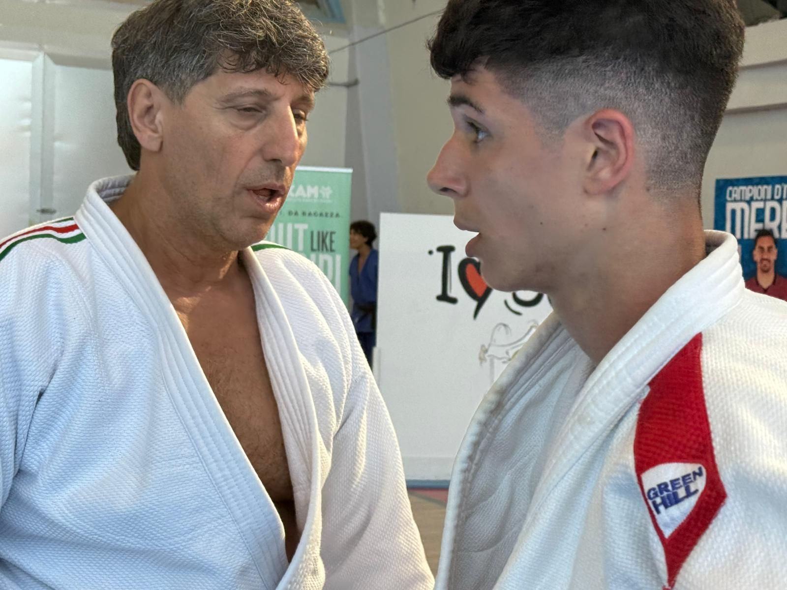 Un uomo in uniforme da judo parla con un ragazzo più giovane, anch'egli in uniforme da judo, in un ambiente chiuso.