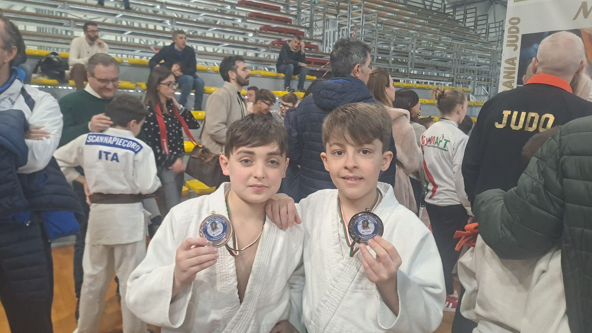 Due ragazzi in uniforme da judo sorridono e tengono in mano le medaglie. Palazzetto dello sport al coperto, spettatori sullo sfondo.