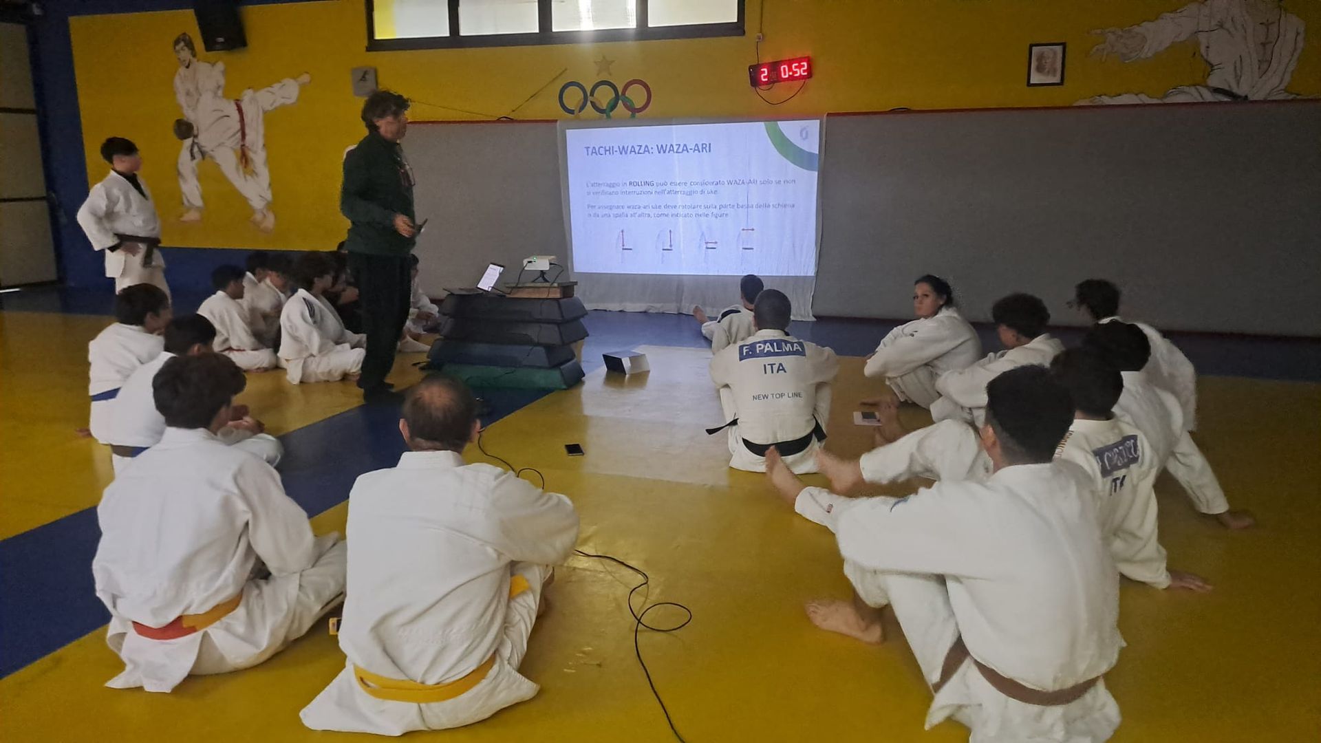Un istruttore tiene una lezione a un gruppo di studenti di judo seduti sul tatami in un dojo. Uno schermo proiettore mostra le informazioni.