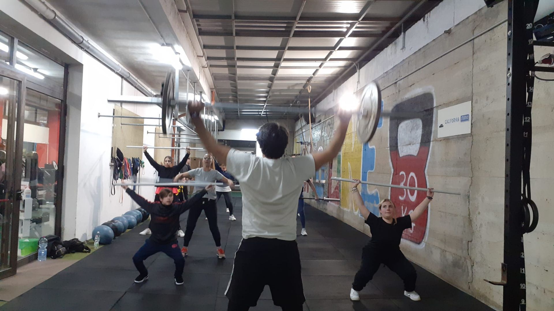Persone che sollevano bilancieri sopra la testa in una palestra. Pareti di cemento, luci fluorescenti e un lungo corridoio.
