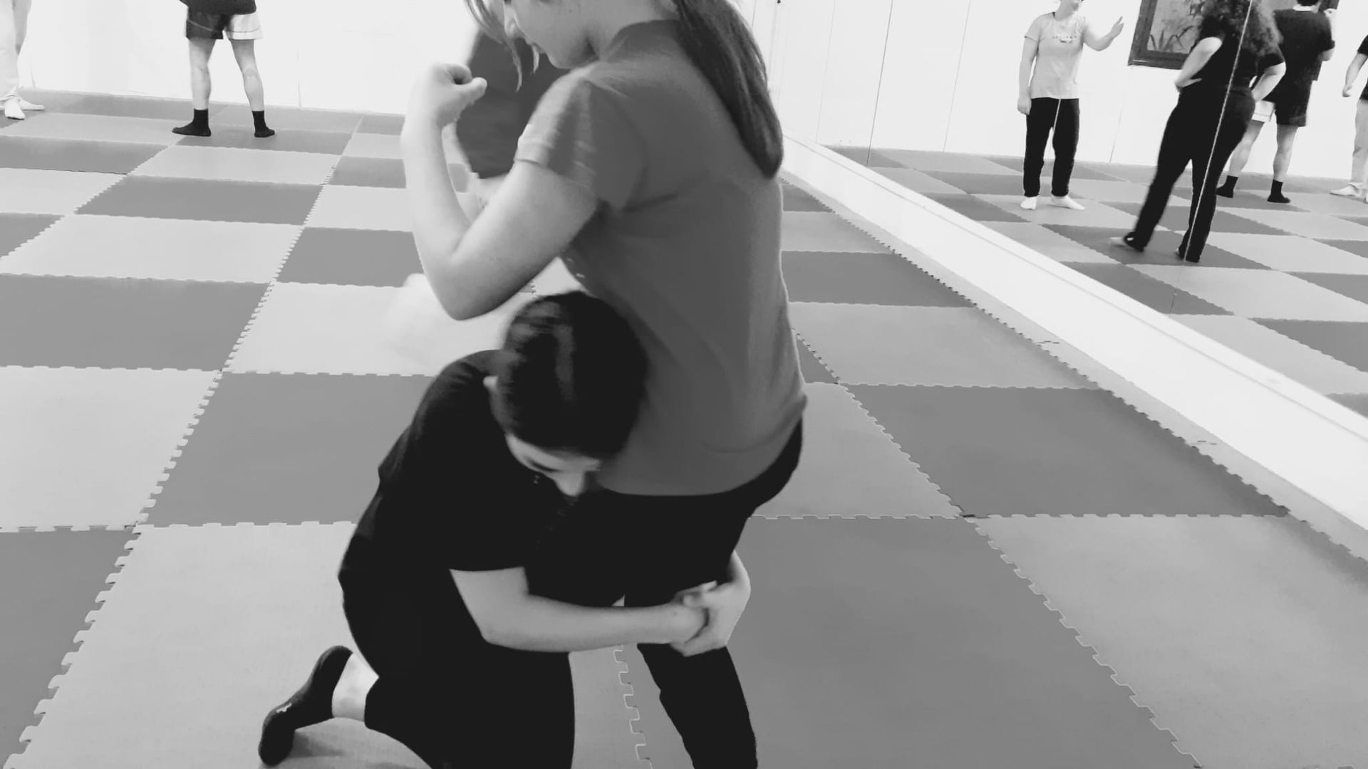 Due persone praticano una mossa di grappling su un pavimento a scacchi in uno studio con uno specchio. Una persona si inginocchia, tenendo le gambe dell'altra.