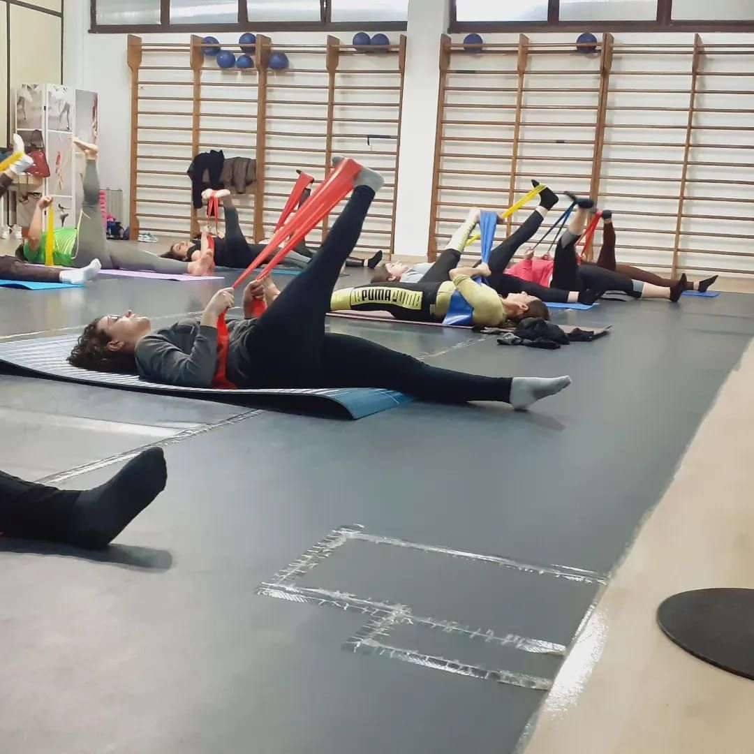 Persone che si allenano sui tappetini in palestra, utilizzando fasce elastiche per allungare le gambe.