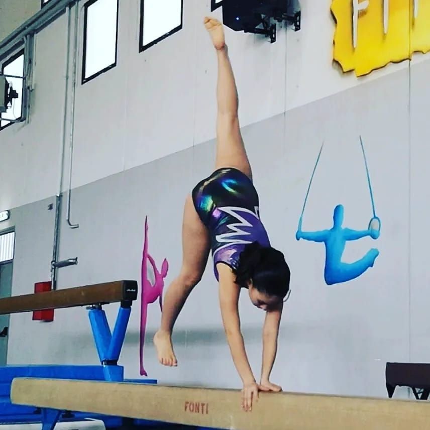 Ginnasta in verticale su una trave di equilibrio, gamba tesa verso l'alto. Ambiente da palestra con attrezzi e decorazioni murali colorate.