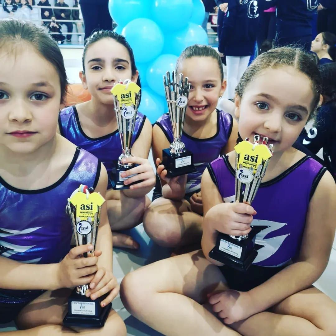 Quattro giovani ginnaste in body viola, con in mano dei trofei, sorridono in una palestra, mentre festeggiano una gara.