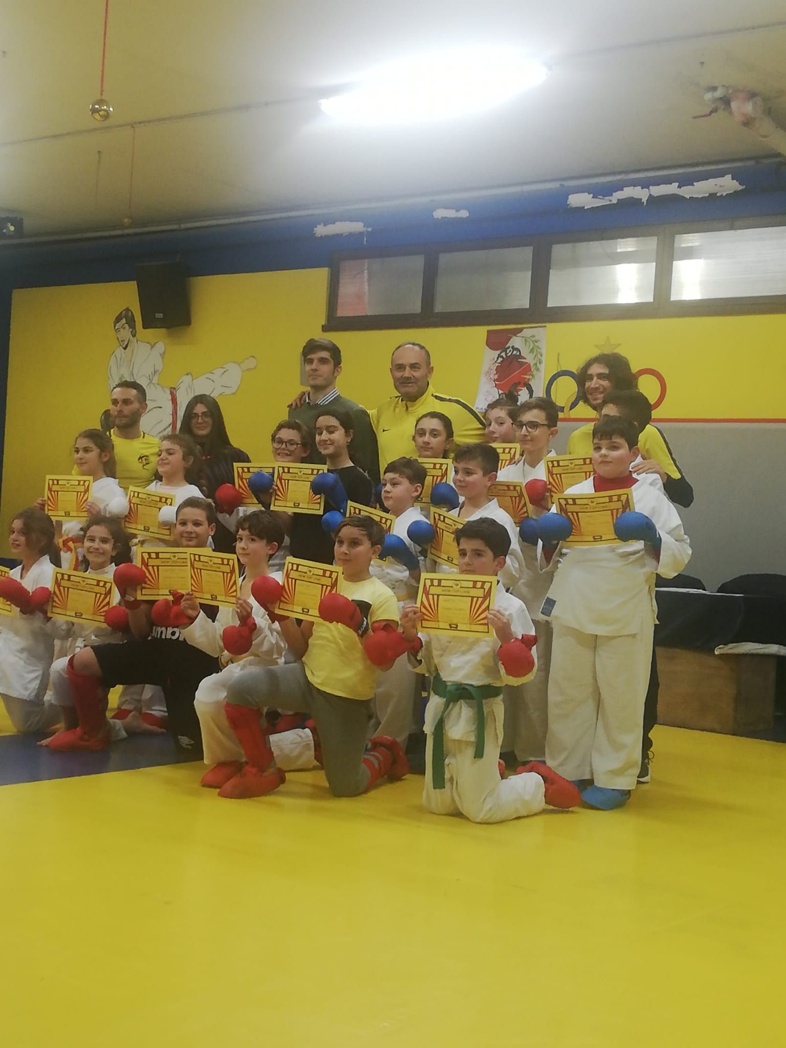 Gruppo di studenti di karate in gi bianchi, con in mano i certificati, in posa su un tappetino giallo, con gli istruttori, in un dojo.