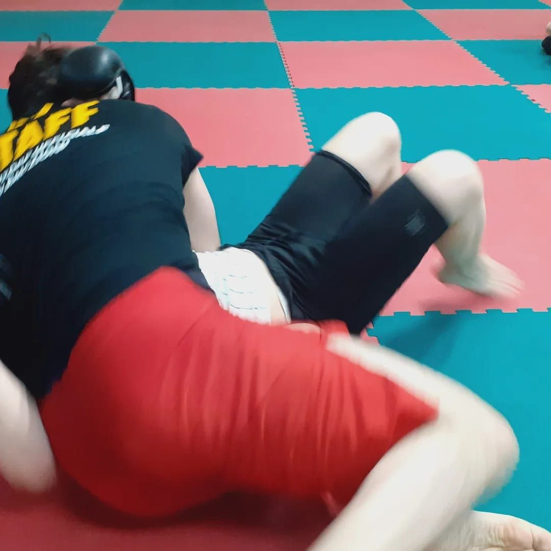 Due persone lottano su un tappetino rosso e verde in una palestra. Una indossa pantaloncini neri, l'altra pantaloncini rossi e una maglietta nera.