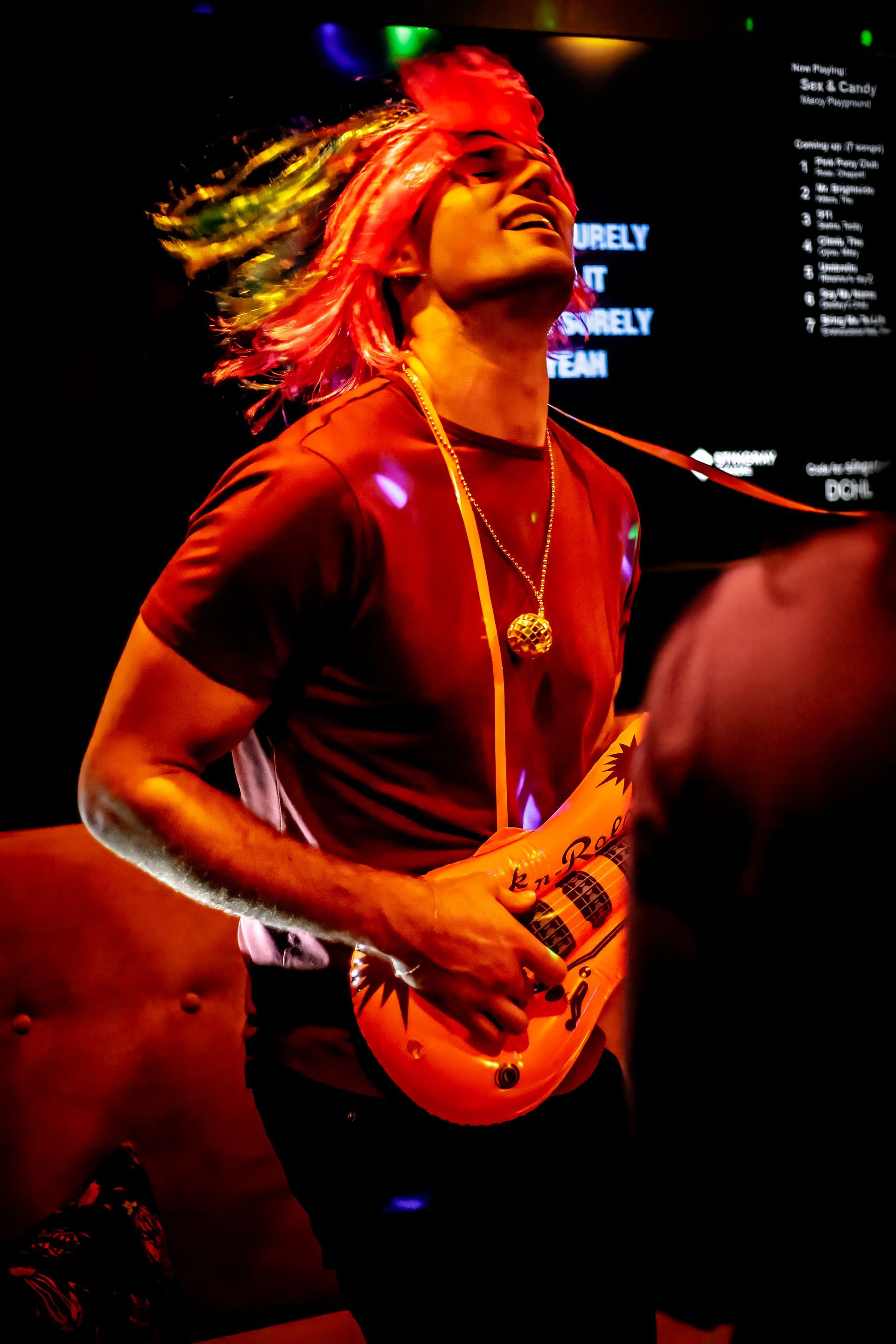 A man wearing a red wig is playing a toy guitar while singing karaoke