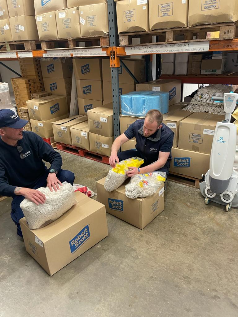 Two men packing items into boxes in a warehouse, surrounded by boxes on shelves.