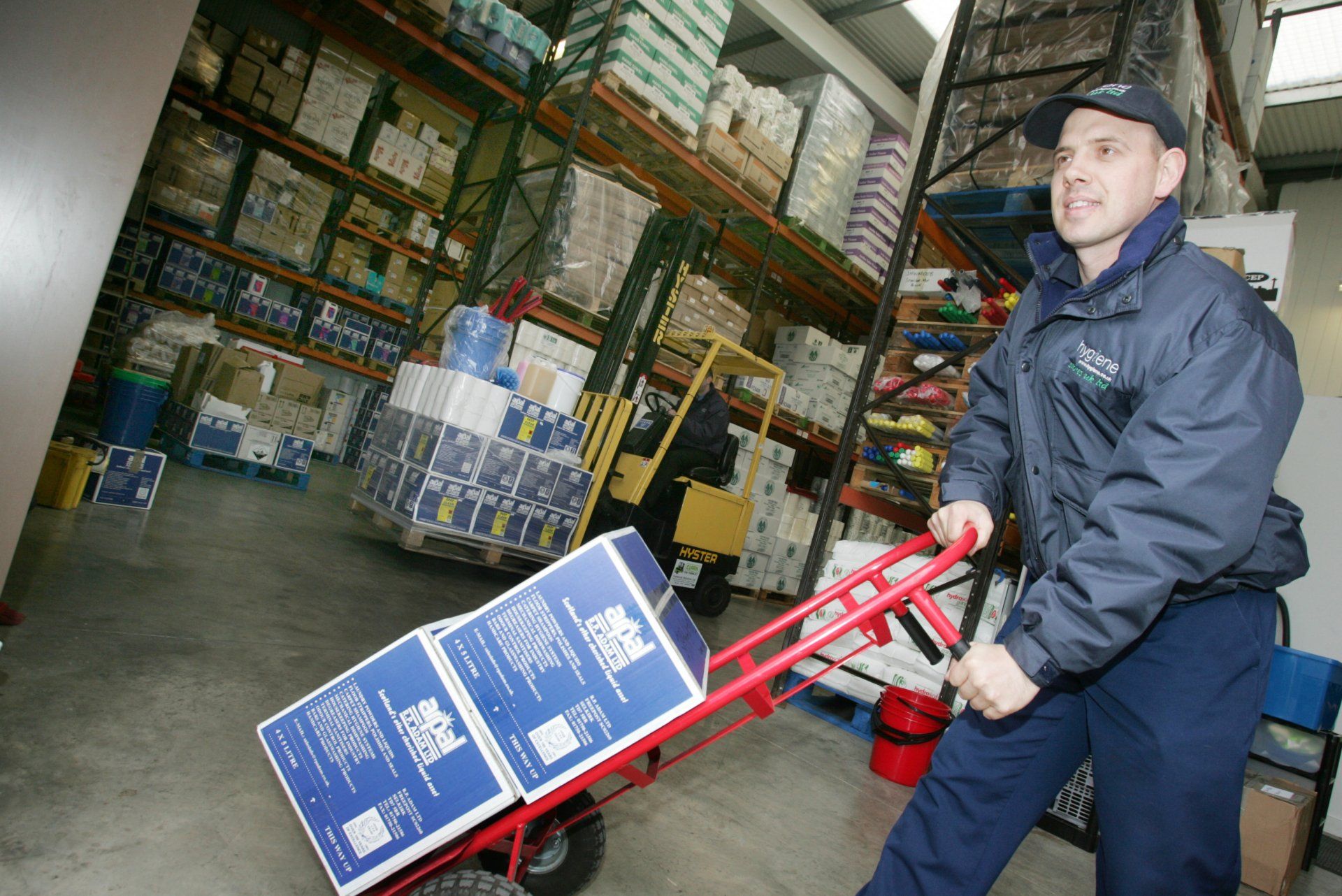 Warehouse worker pulling a hand truck loaded with boxes. Shelves of goods and a forklift in the background.
