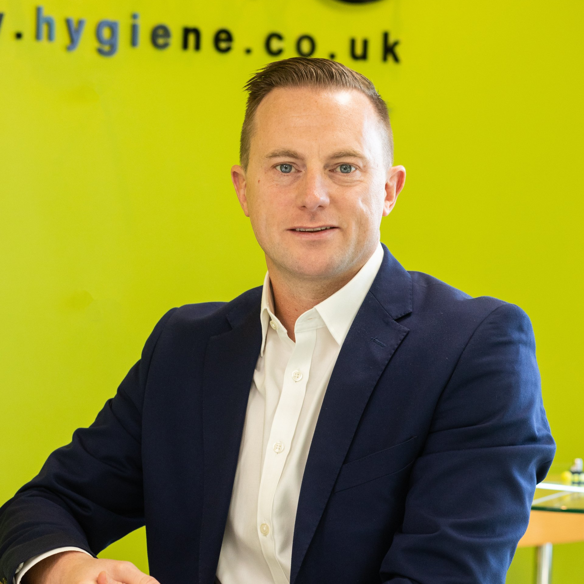 Man in a navy blazer and white shirt, smiling, against a green background.