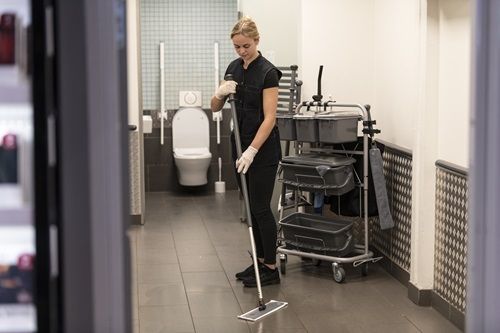Woman in black uniform mops a tiled floor near a cleaning cart and restroom.