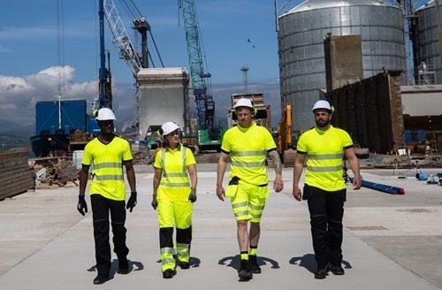 Four construction workers in neon yellow walking toward the camera. Background shows cranes and industrial buildings.