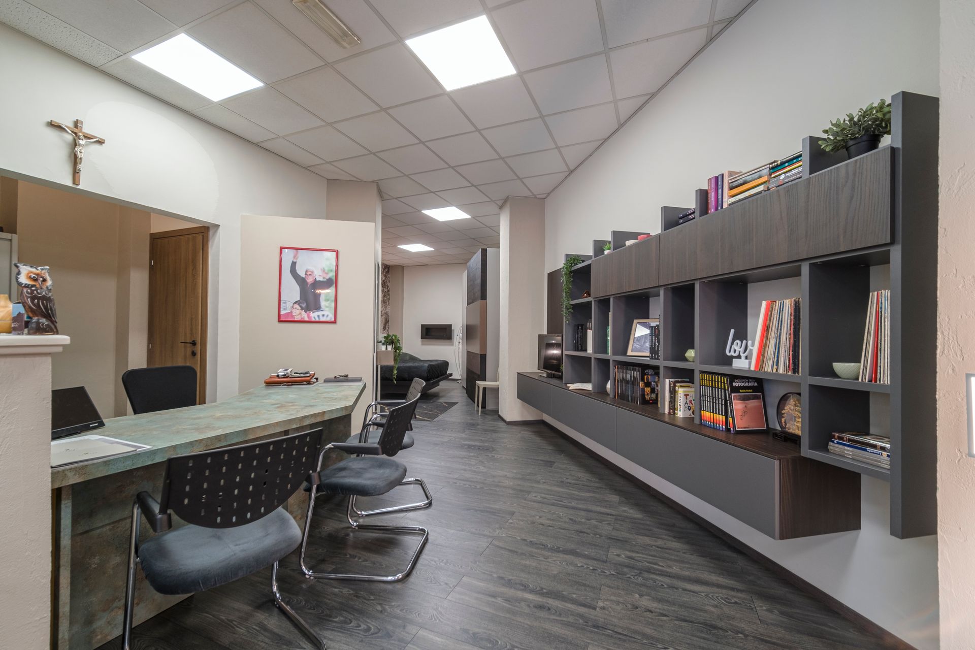 Un lungo corridoio con un tavolo, sedie, scaffali e una croce sul muro.