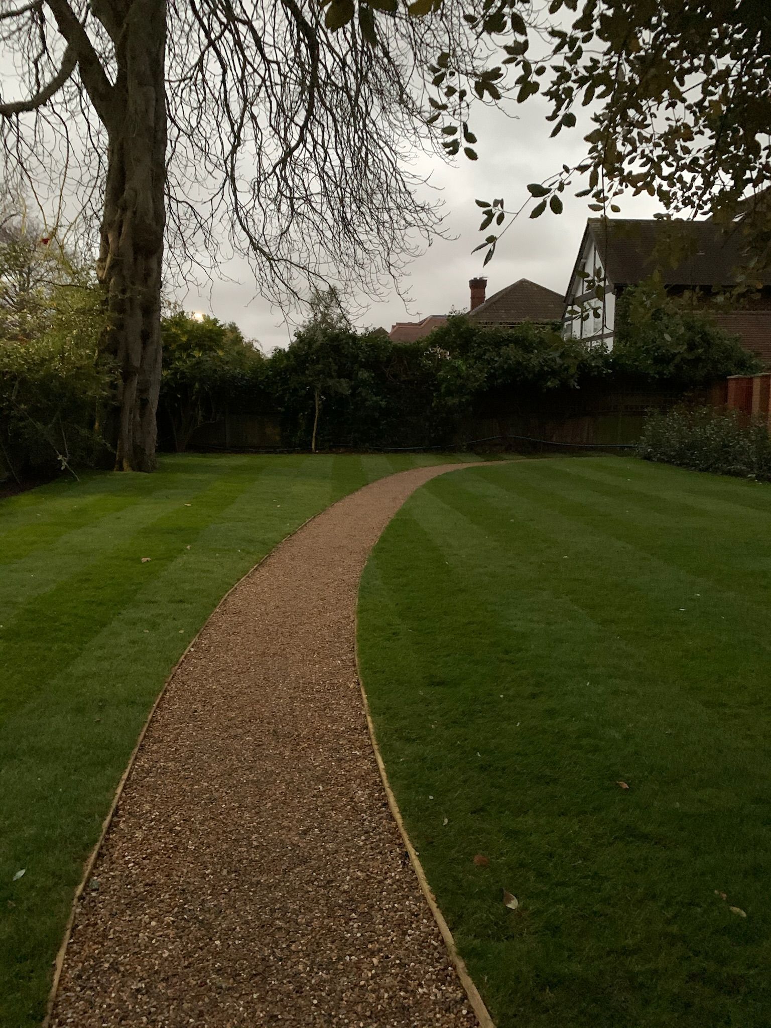 Gravel path curves through a manicured lawn, leading toward trees and houses under an overcast sky.