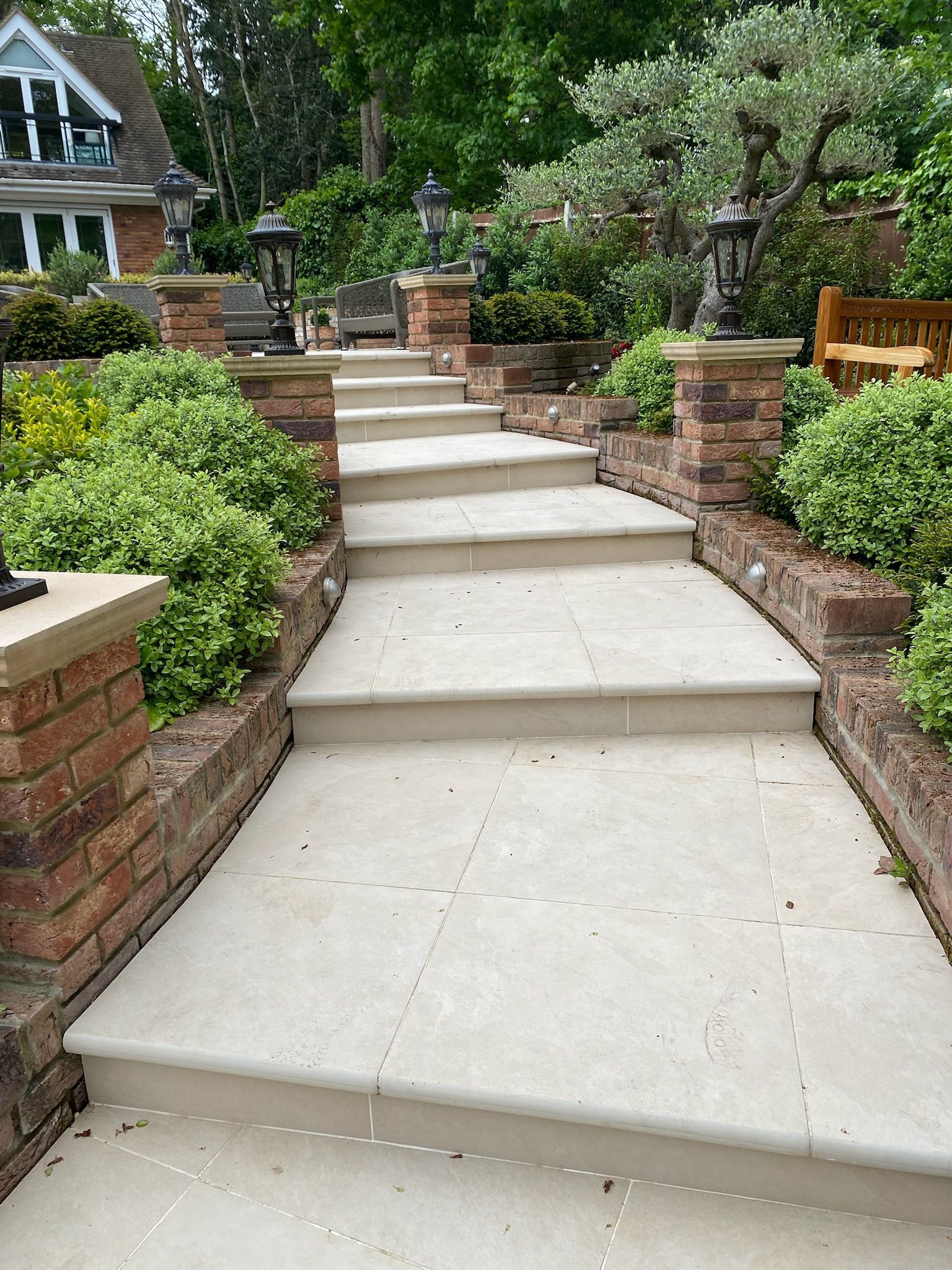 Stone steps leading up a garden path, flanked by brick walls, bushes, and decorative lights.