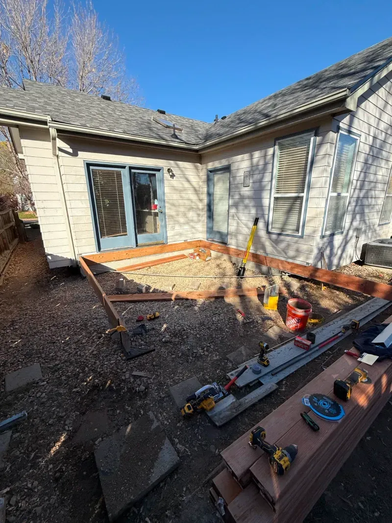 Backyard deck under construction; tools and materials scattered. Brown deck boards, tan house, blue sky.