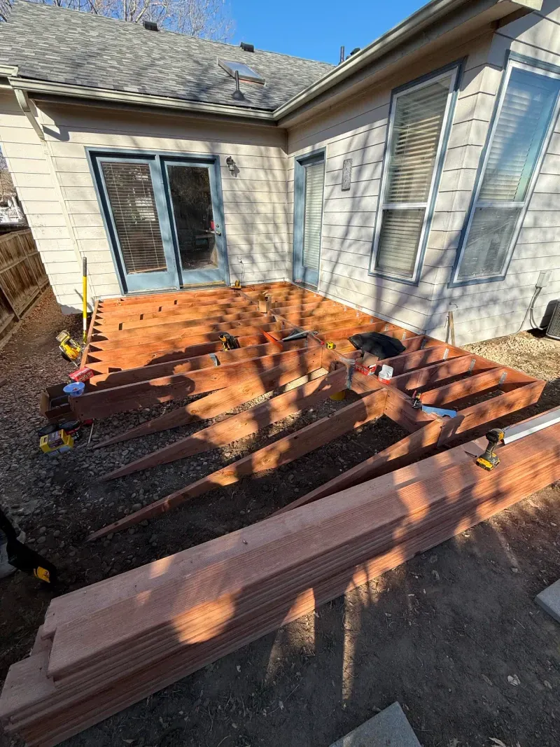 A partially built deck with reddish-brown wood.  Tools and wood planks are scattered around the structure outside a home.