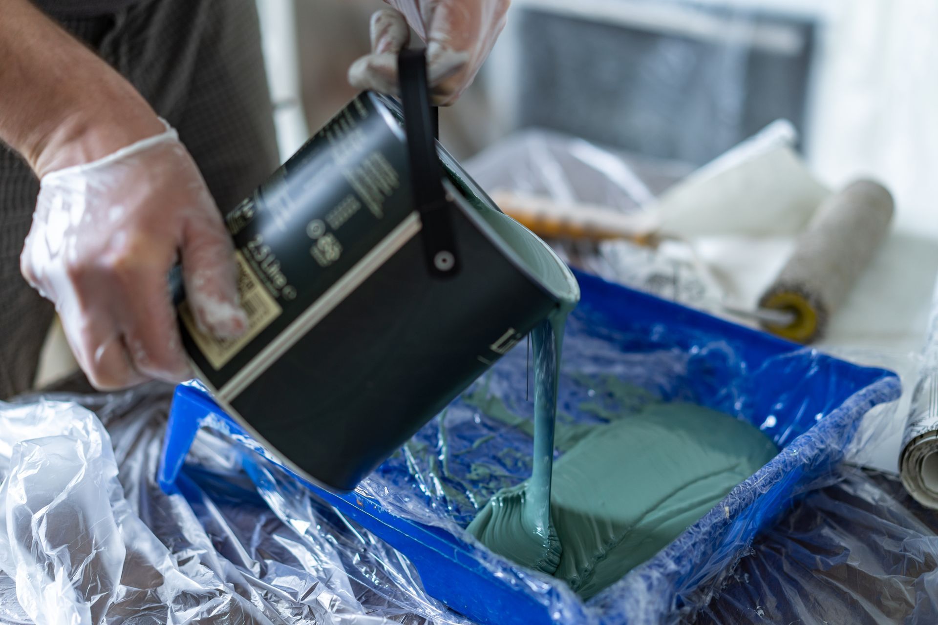Person pouring teal paint from a can into a blue paint tray.