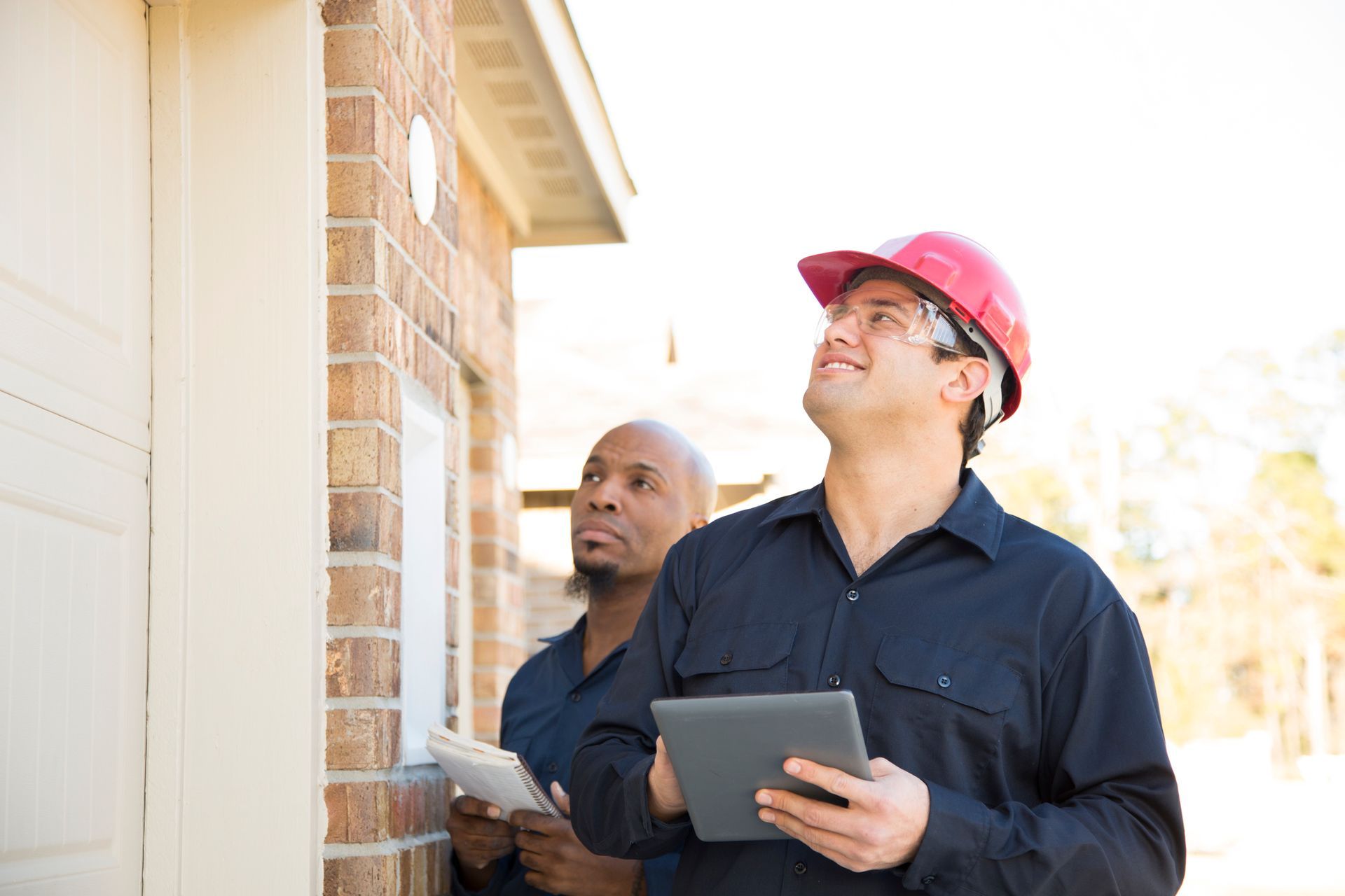 Two construction workers are looking at a house while one is holding a tablet.