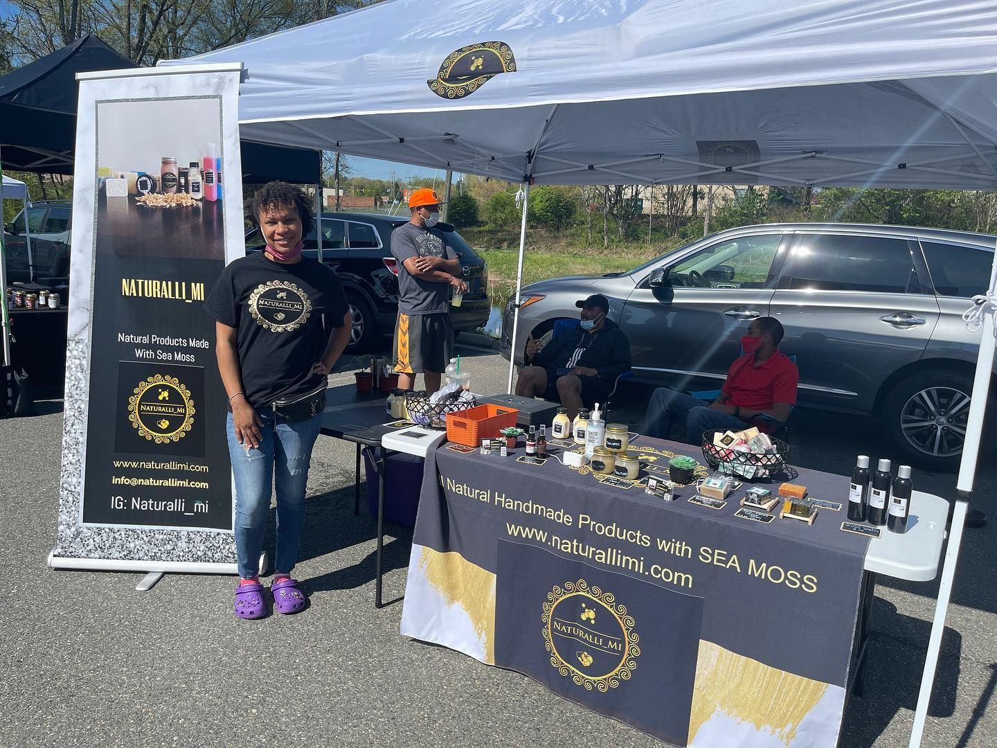 Woman at a booth selling products labeled "Wellness with Sea Moss". Others nearby. Sunny outdoor setting.