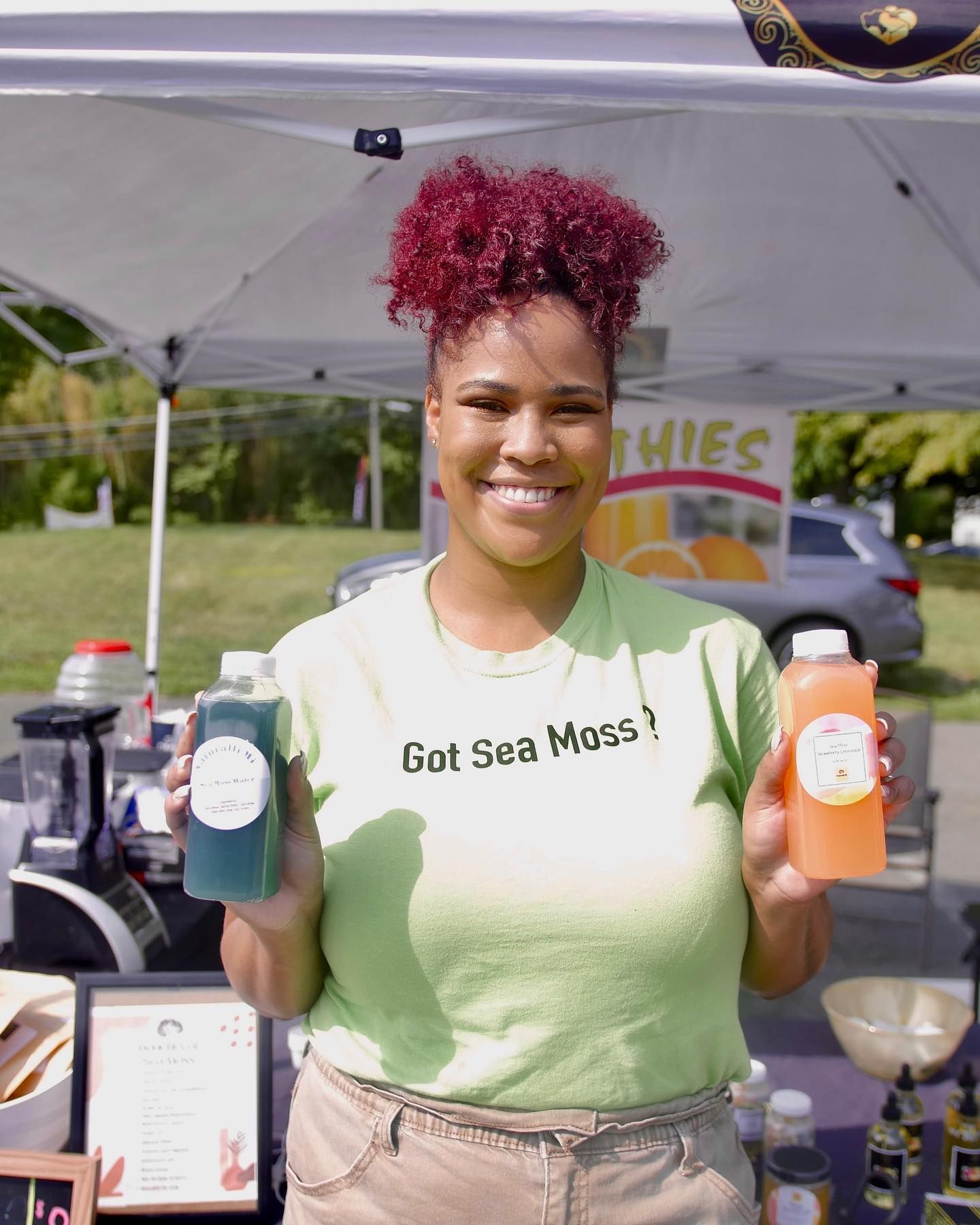 Woman with red hair holds two bottles of Sea Moss smoothies at an outdoor market, smiling.