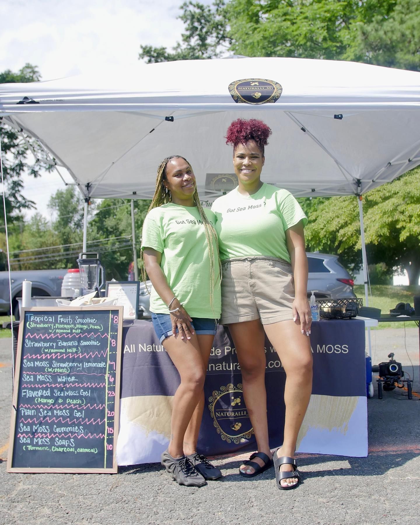 Two women stand at a market stall, wearing green shirts. One has braids; the other, curly red hair.
