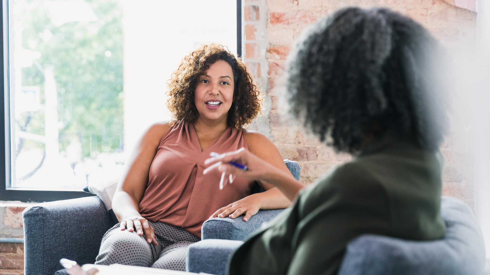 Woman in therapy session with therapist. Both seated in armchairs; therapist gestures while talking.