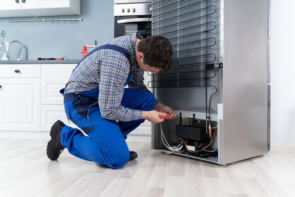 A repair technician in blue overalls is working on the back of a refrigerator in a kitchen.